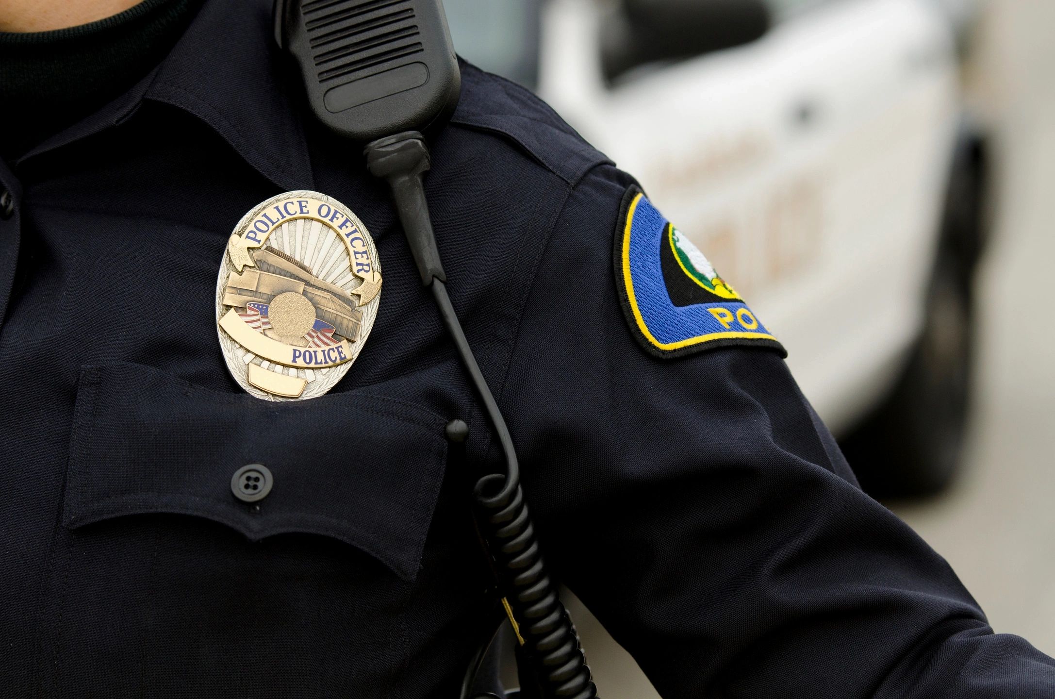 Close-up of a police officer in a uniform, highlighting the metallic chest badge and shoulder-mounted radio microphone.